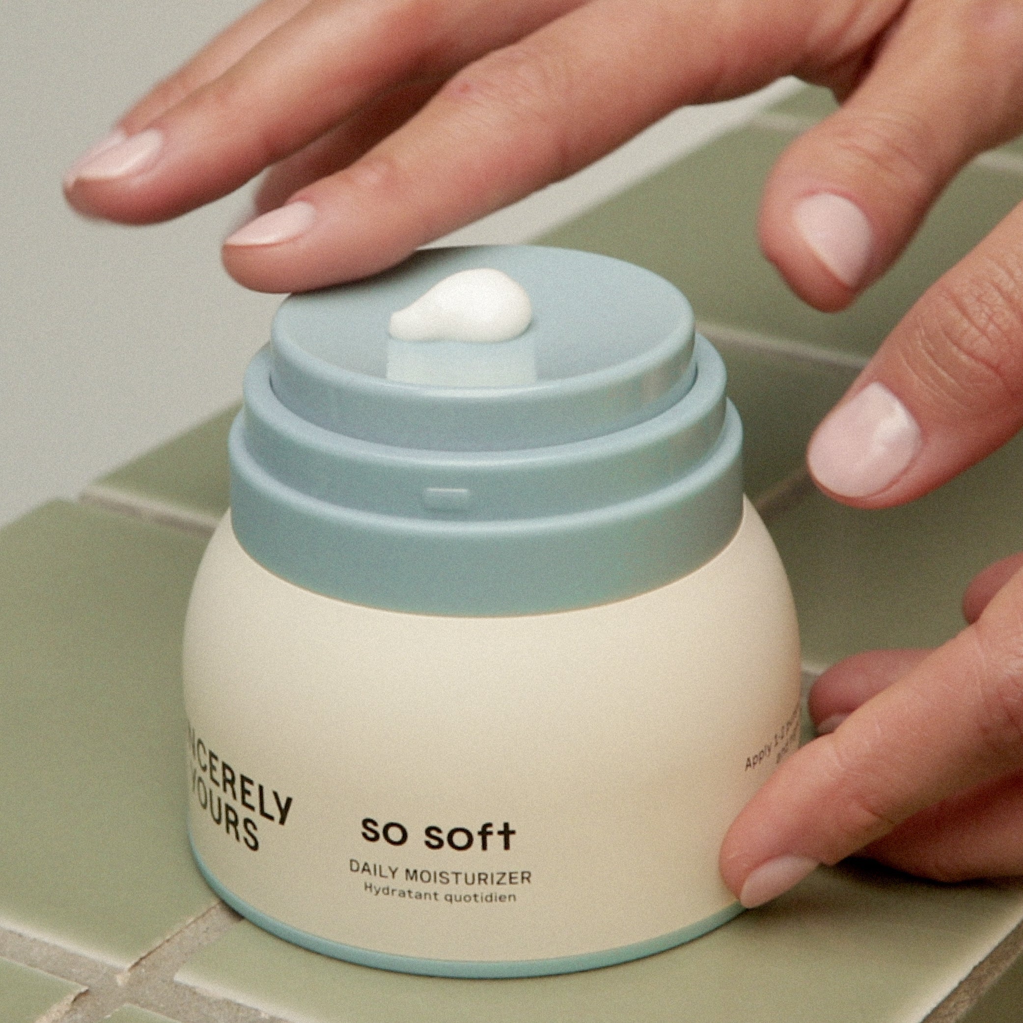 Hand applying cream from a jar labeled 'So Soft' on a tiled surface