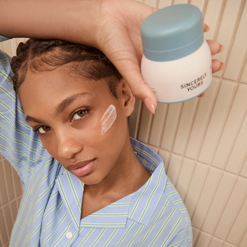 Person applying cream to face with 'Sincerely Yours' jar in hand, tiled bathroom background